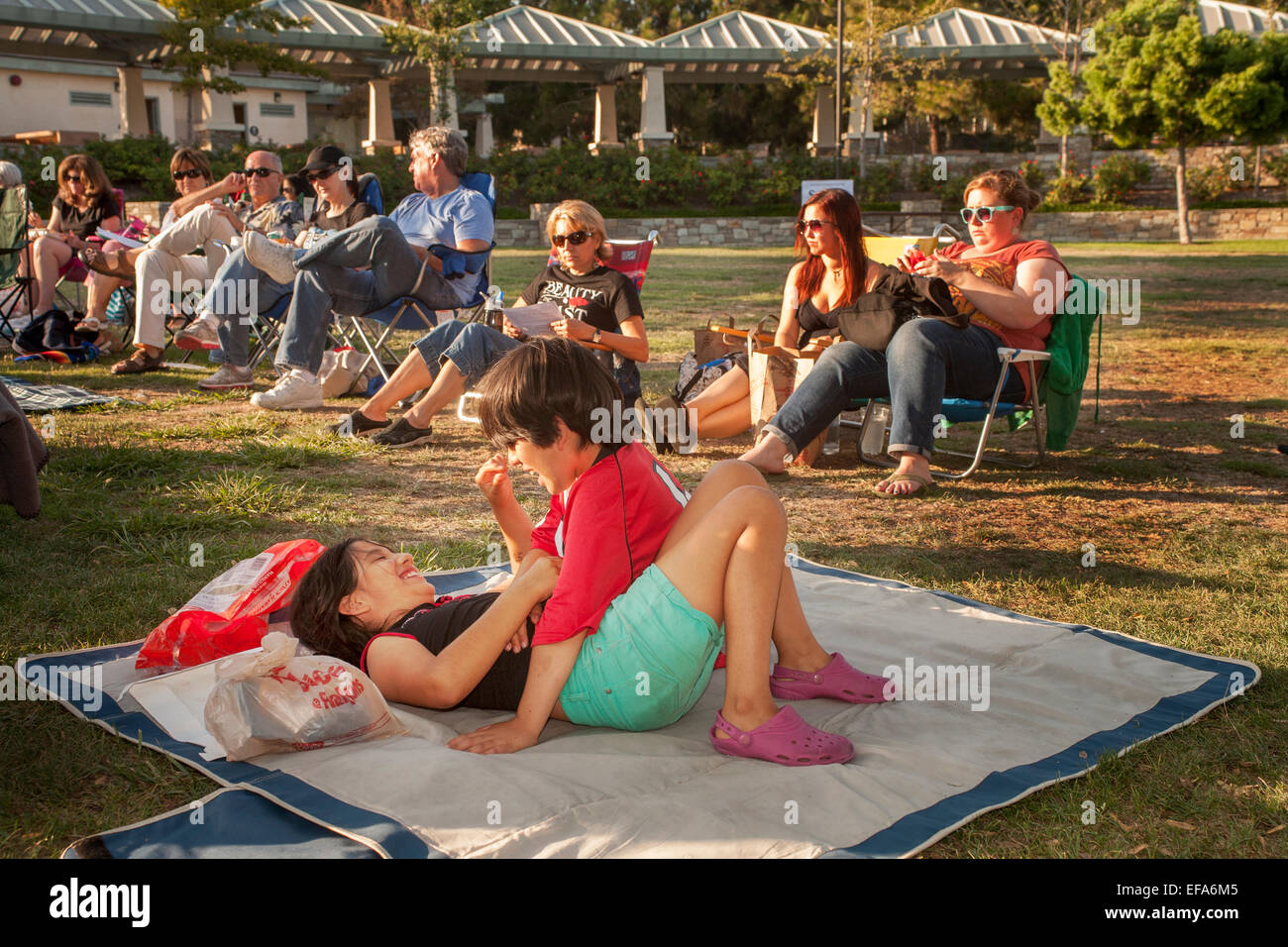 En el sol de la tarde, un hermano y hermana Hispana juega sobre una manta en un Laguna Hills, CA, Parque. Nota Los adultos en segundo plano. Foto de stock