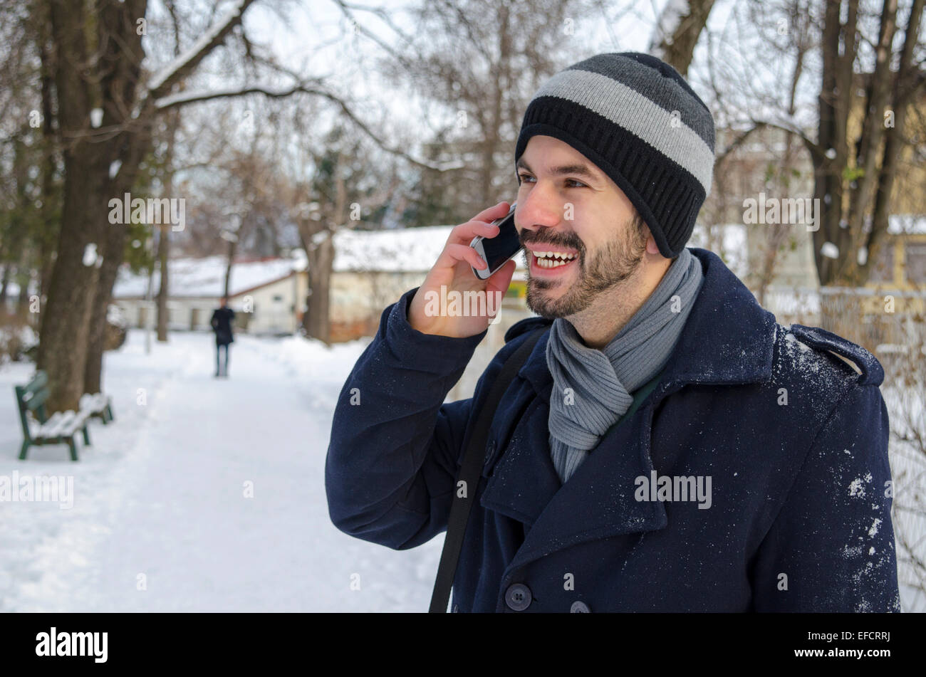 Macho joven barbudo hablando por su teléfono inteligente, afuera en la nieve. Foto de stock