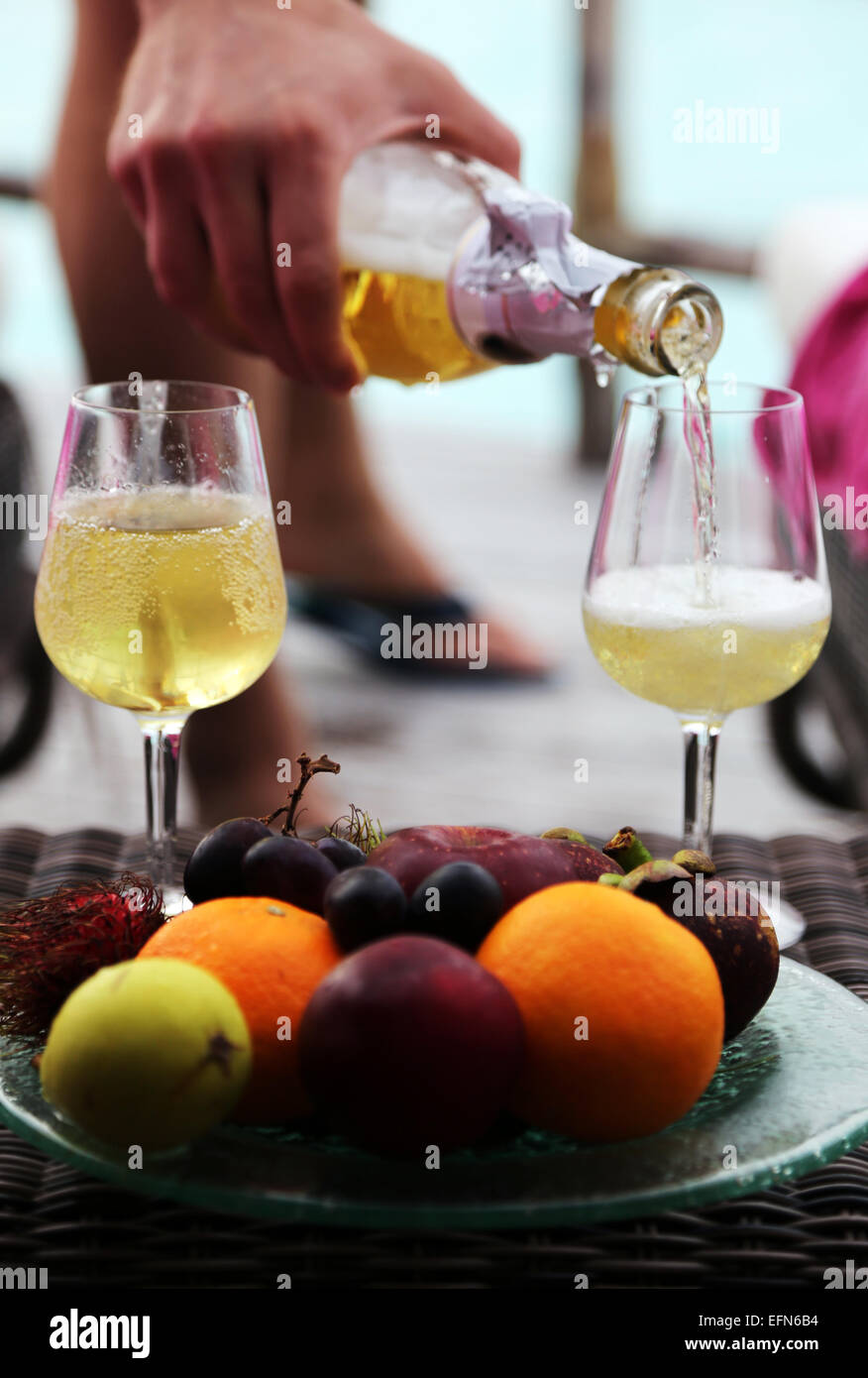 Una mano de hombre vierte en dos copas de champán. Un plato de frutas exóticas está en el frente. Foto de stock