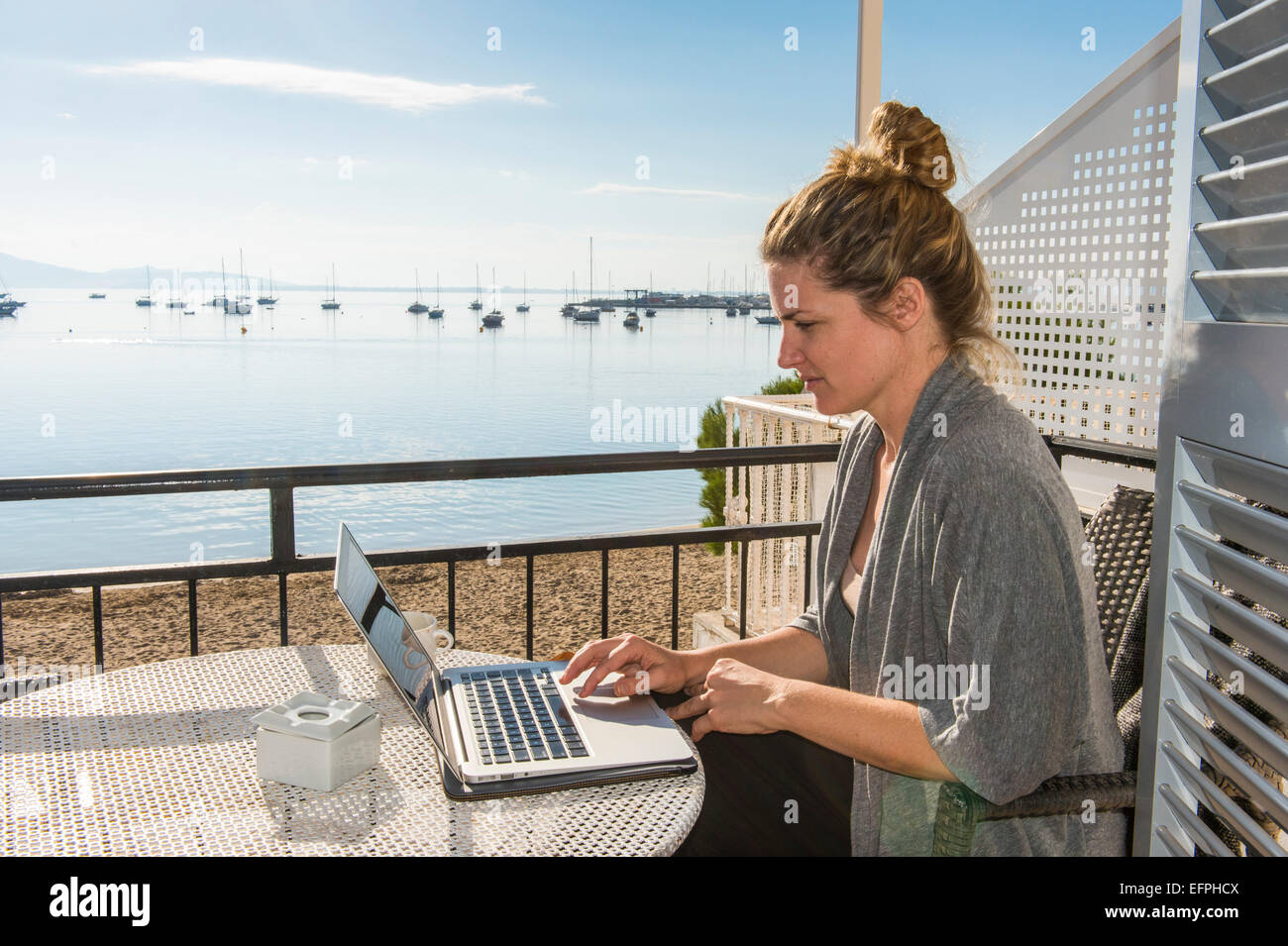 Mujer trabajando en su laptop en un balcón con vistas al océano, Port de Pollença, Mallorca, Islas Baleares, España Foto de stock