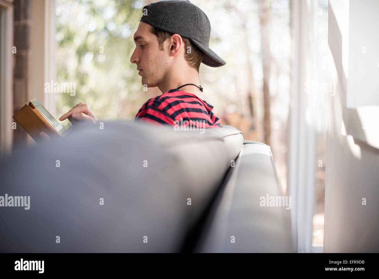 Hombre que llevaba una gorra de béisbol hacia atrás, sentado en un sofá, mirando a una tableta digital. Foto de stock