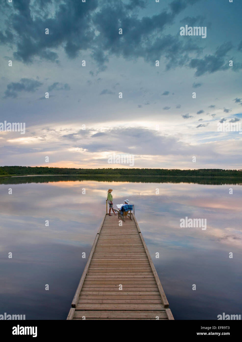 Una pareja, hombre y mujer sentada al final de un largo muelle de madera llegando a un lago tranquilo, al atardecer. Foto de stock