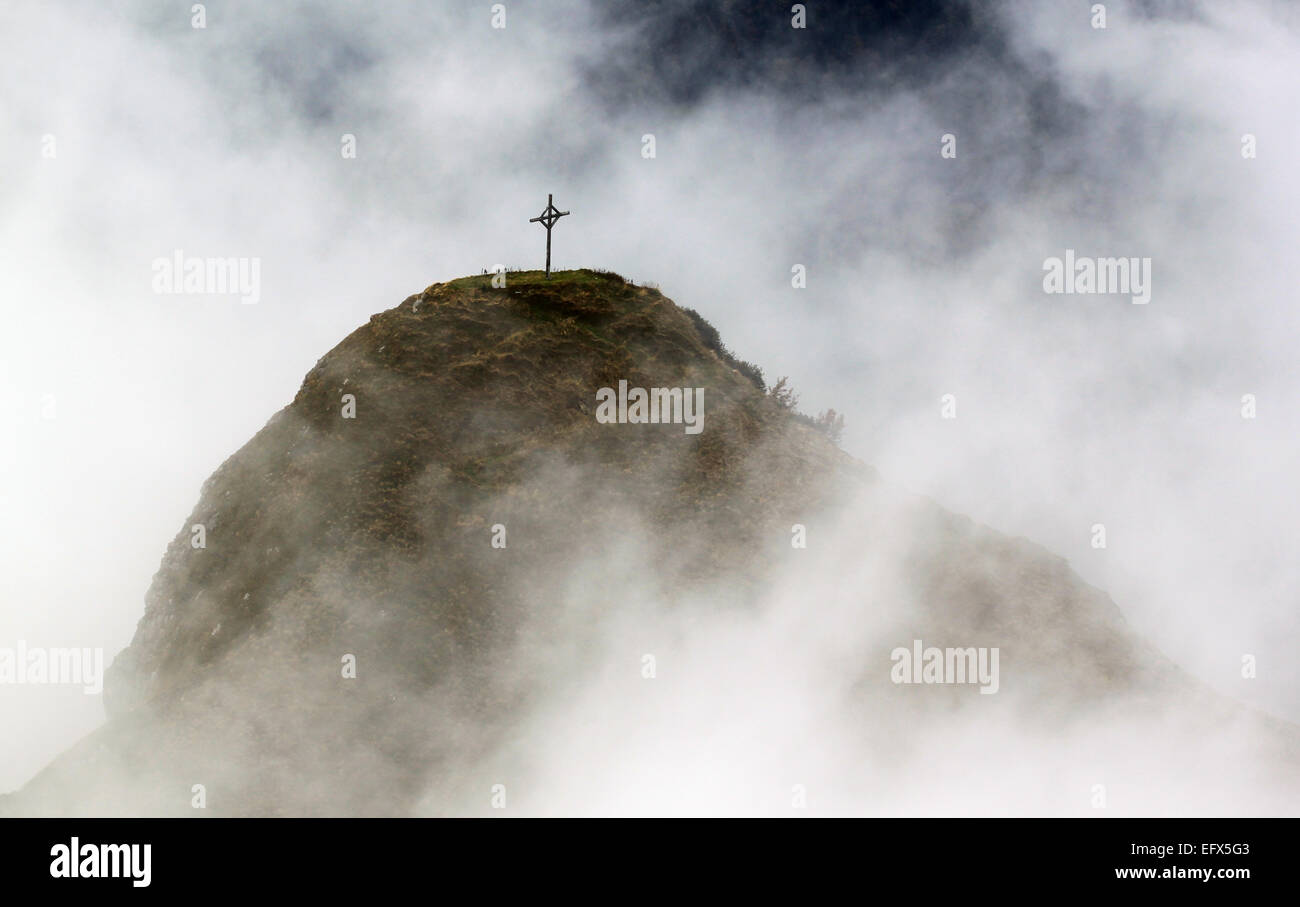Una sencilla cruz se alza sobre un Mountain envuelta en nubes blancas. Foto de stock