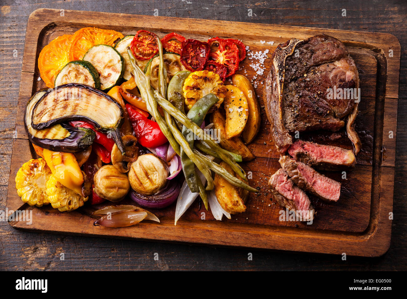 Club de bistec de carne con salsa de pimienta y verduras asadas en placa de corte sobre fondo de madera oscura. Foto de stock