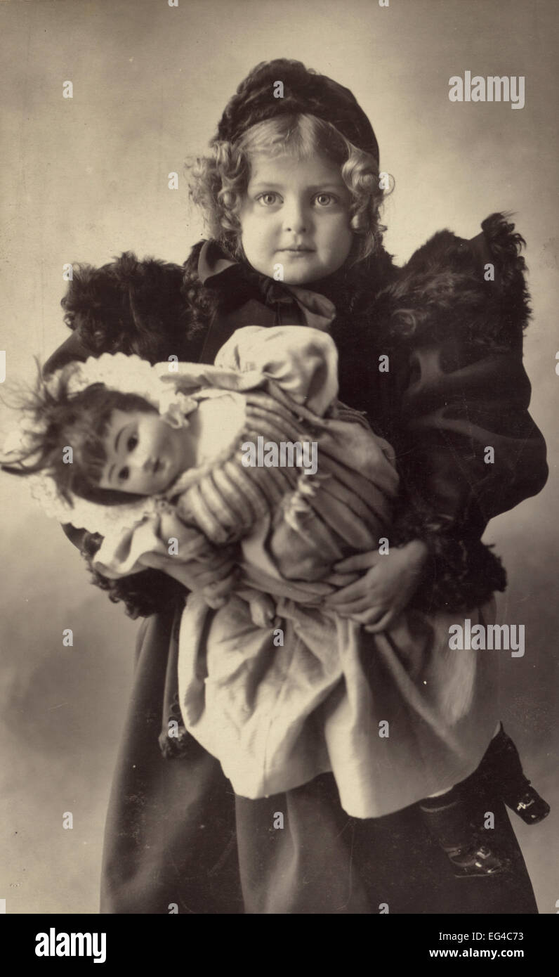 Una chica joven vestida con un abrigo de pieles recortada y sombrero, llevando su muñeca, circa 1898 Foto de stock