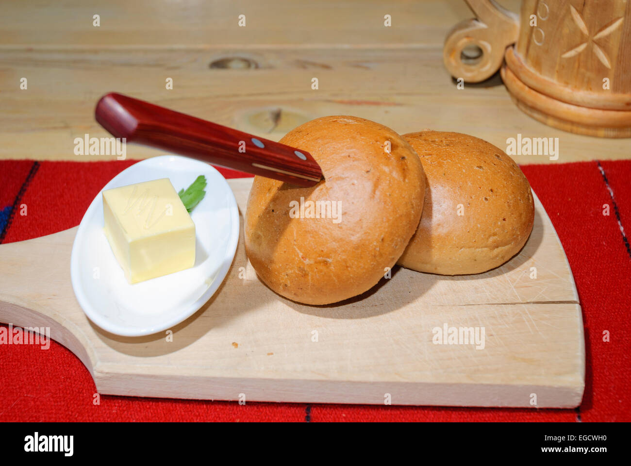 Comida tradicional Baltic - rollos de pan y una perilla de mantequilla sobre una tabla de cortar de madera, con un flagon de cerveza en el fondo Foto de stock