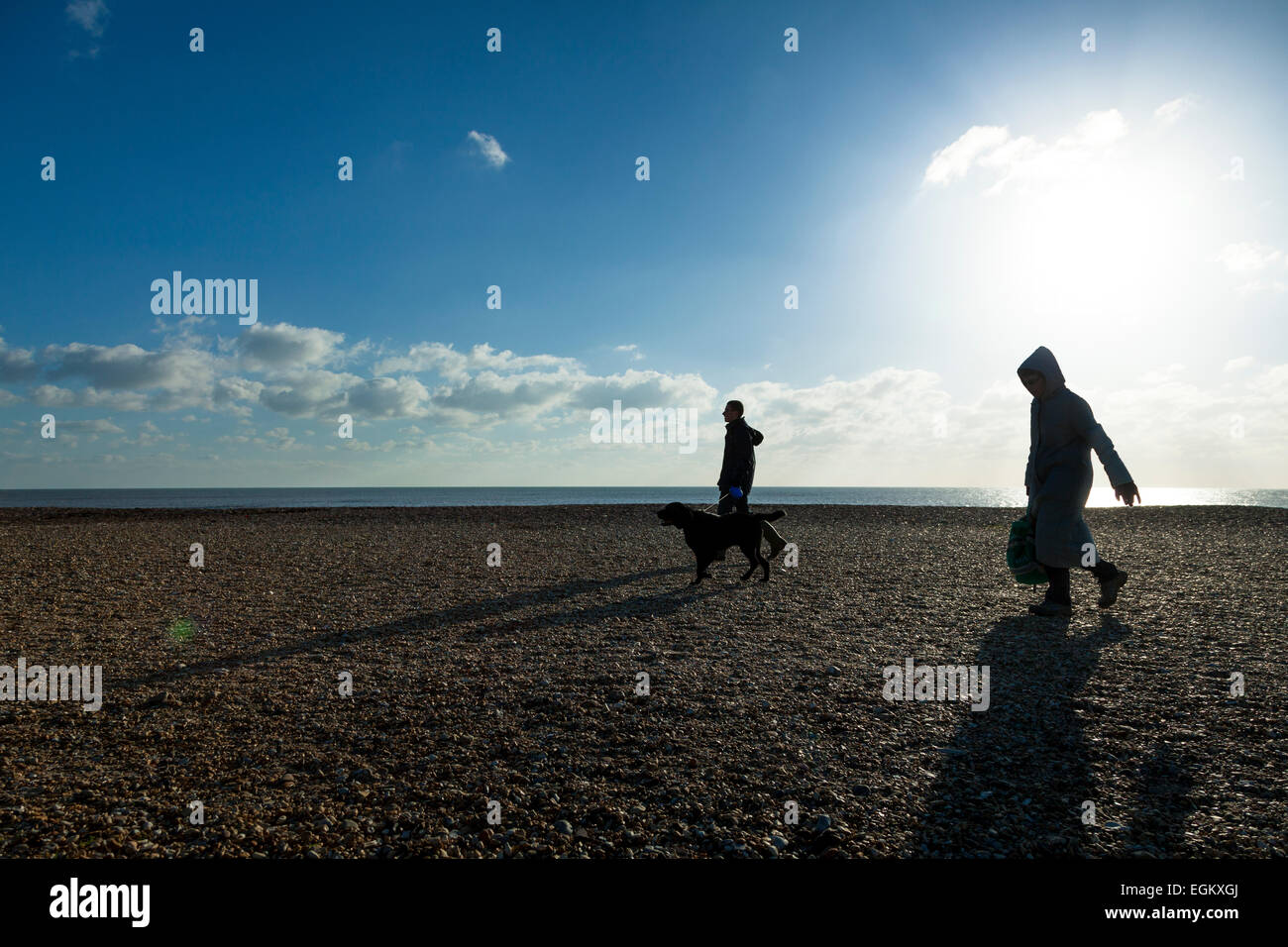 Dos personas caminando un perro en una playa de guijarros en un soleado día de invierno con sun flare Foto de stock