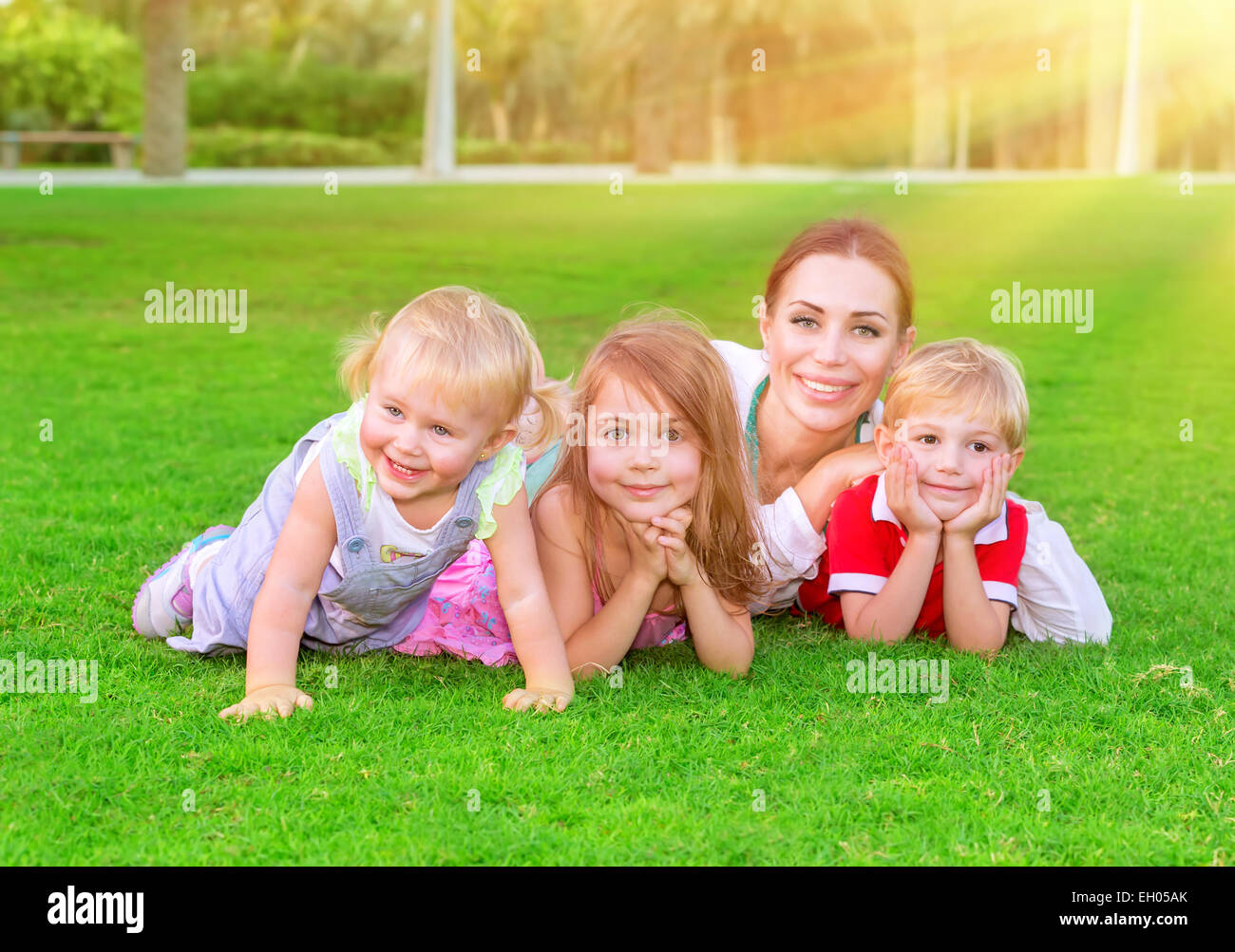 Feliz familia divirtiéndose al aire libre, hermosa madre con tres cute little children acostada en la hierba verde fresca Foto de stock