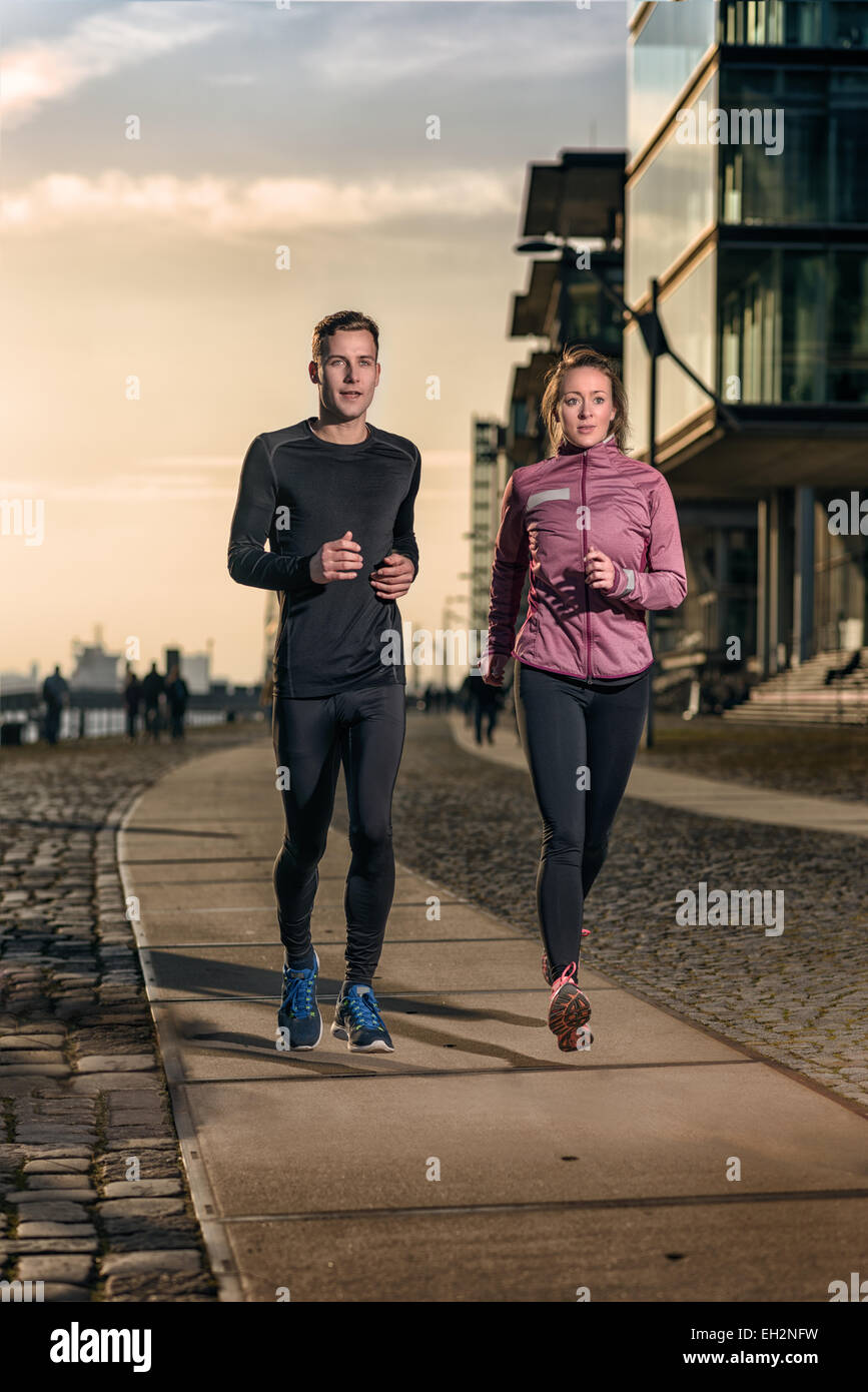 Active pareja joven correr al lado de la otra en el paseo marítimo al atardecer durante su entrenamiento diario en la salud y el fitness conc Foto de stock