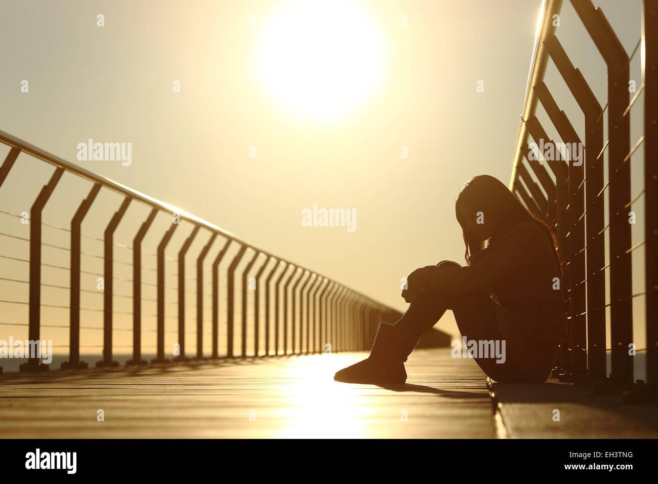 Triste joven deprimido sentado en el piso de un puente en la playa en el atardecer. Foto de stock