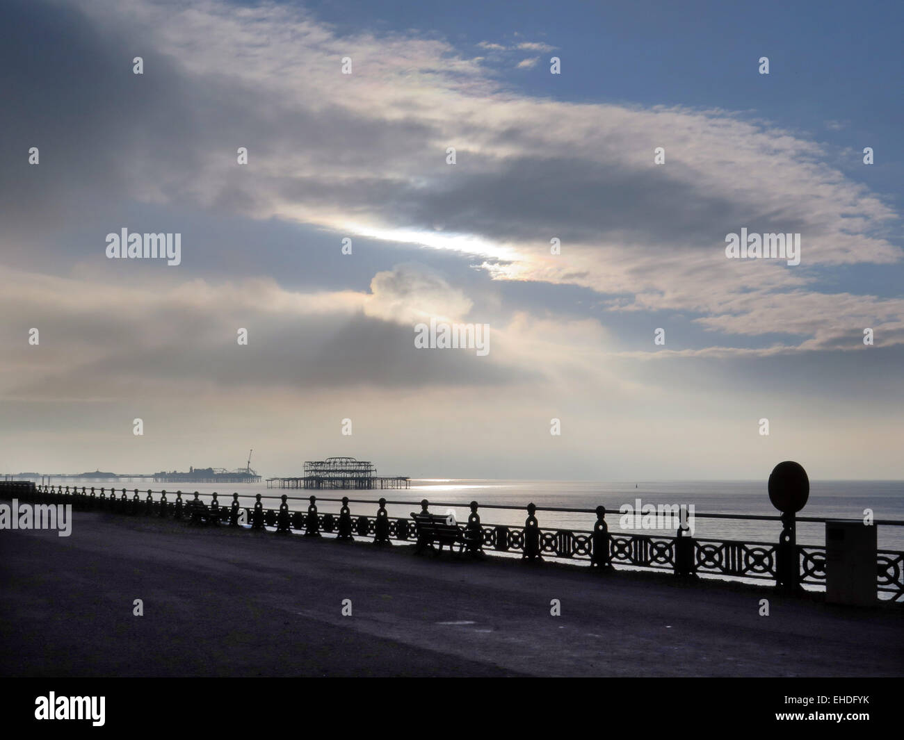 El incendio, atacó a la ruina del West Pier, Brighton diseñado por Eugenius Birch, visto a través de la promenade con pasamanos decorativos y bancos a un brillante mar y nubes cirrus Foto de stock
