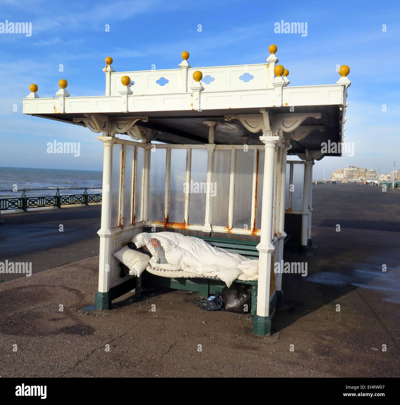 Un hombre sin hogar disfruta de un cigarrillo mientras envuelto en un saco de dormir del frío en un paseo marítimo de estilo Eduardiano cobijo en Hove promenade, en la ciudad de Brighton y Hove, East Sussex Foto de stock