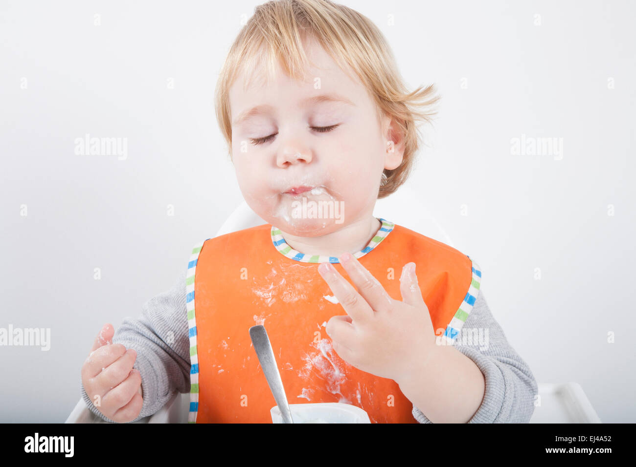 Rubio caucásico diecisiete meses de edad del bebé babero naranja suéter gris comiendo comida con su mano en la silla alta blanco Foto de stock