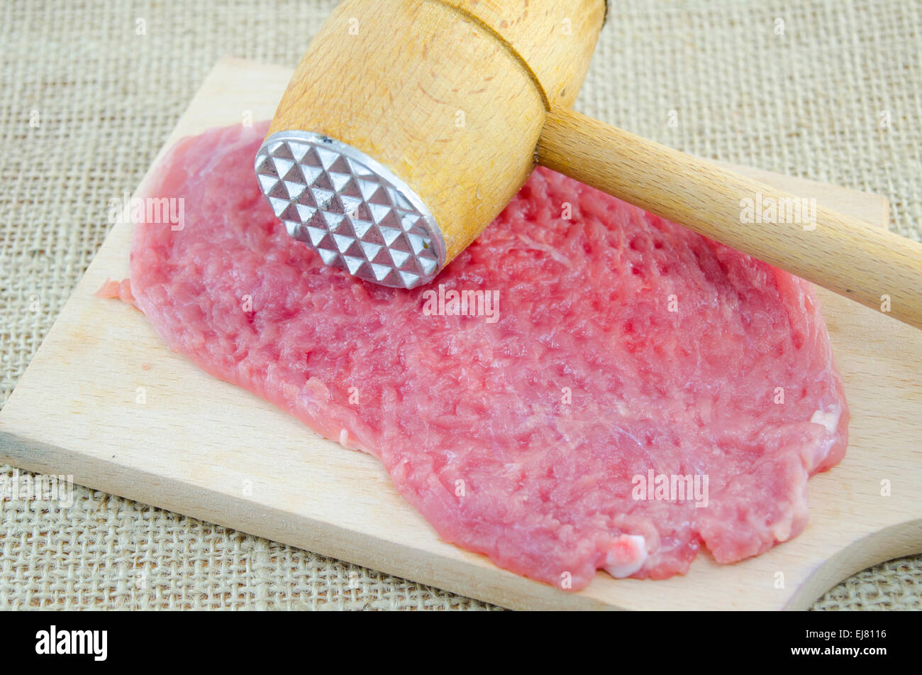Raw bistec de carne roja y un martillo sobre una tabla para cortar madera. Colocados sobre una mesa cubierta con un mantel vintage Foto de stock