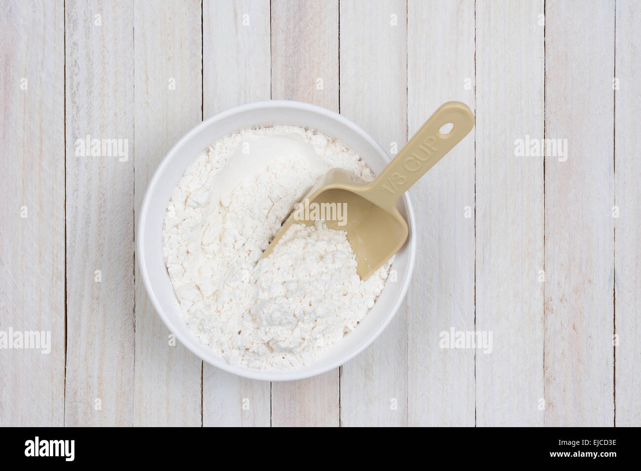 Imagen aérea de un tazón de harina sobre una mesa de cocina rústica de madera blanca. Una cuchara de plástico está atascada en la harina. Foto de stock
