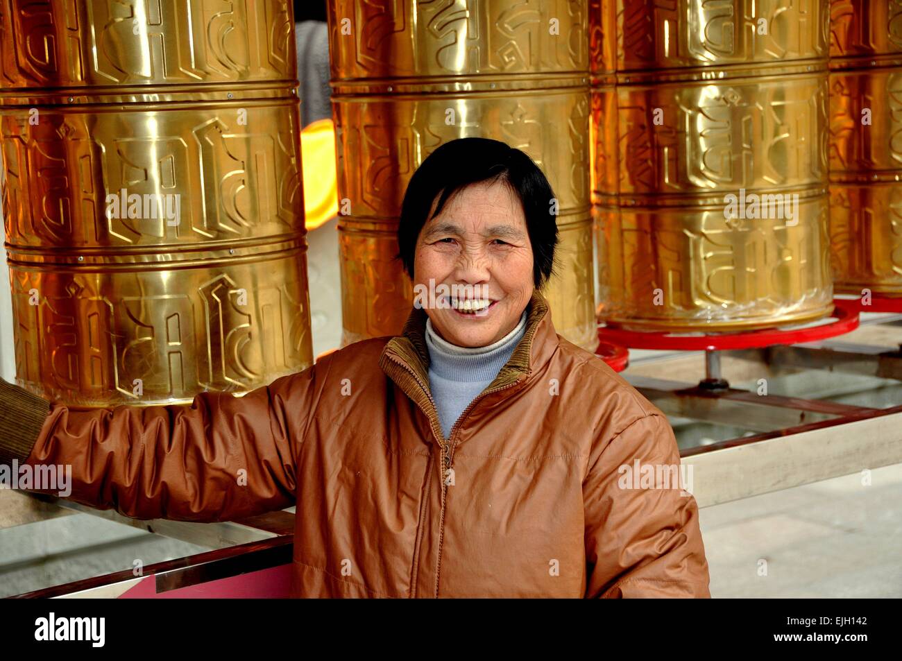 Pengzhou, China: China sonriente mujer de pie junto a una fila de estaño rueda de oración tibetano de tambores en el templo Xing largo Foto de stock
