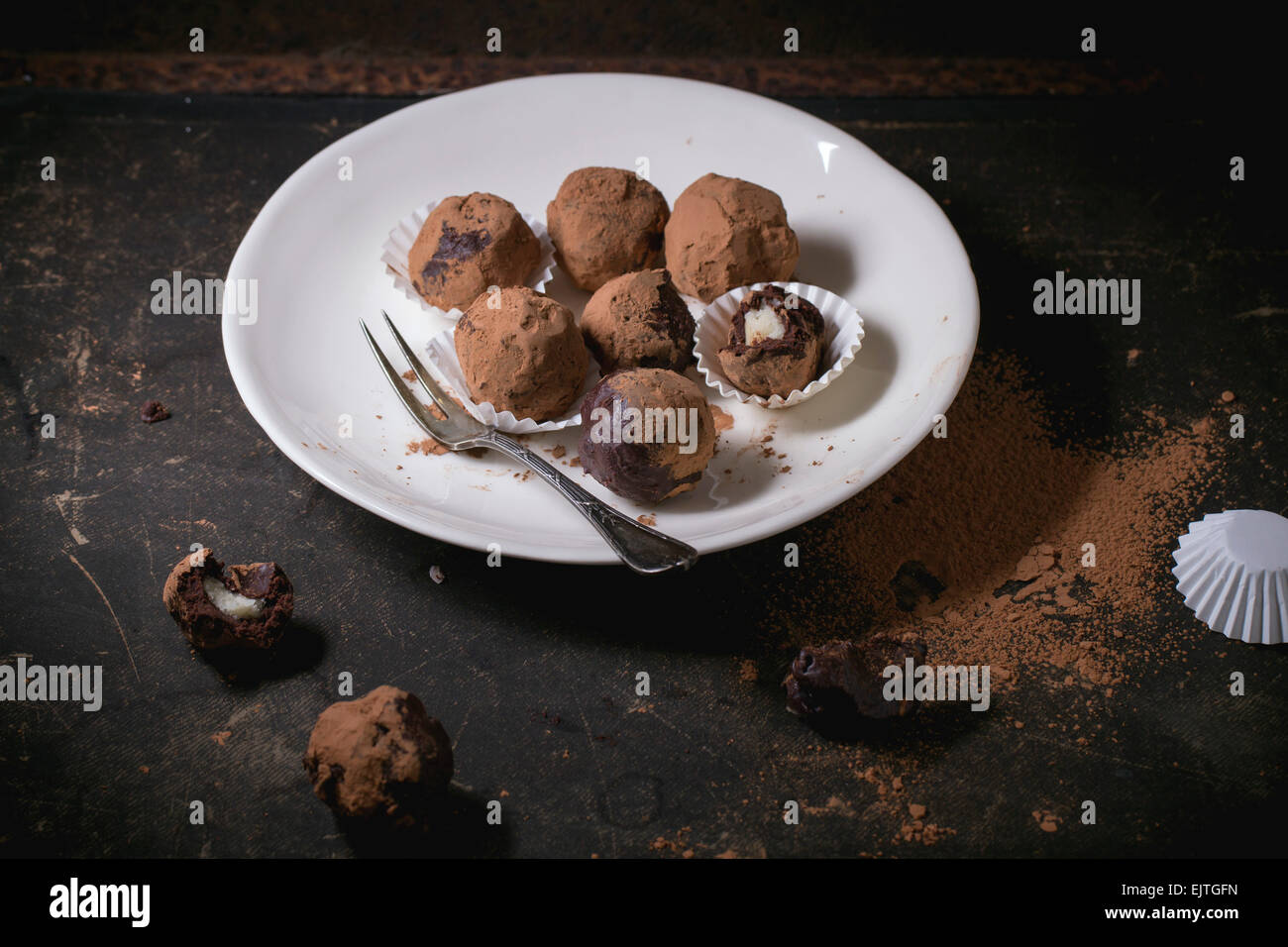 Placa blanca con trufas de chocolate caseras con mazapán y cacao en polvo sobre tabla oscura. Foto de stock