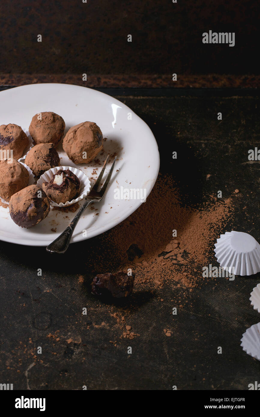 Placa blanca con trufas de chocolate caseras con mazapán y cacao en polvo sobre tabla oscura. Foto de stock