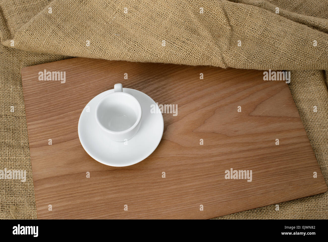 Taza de café blanco vacío sobre una placa de madera con tela de fondo. Foto de stock