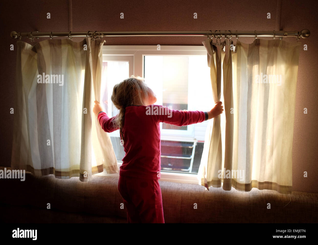 Un soñoliento joven en pijama abre las cortinas para mirar fuera de la ventana en el inicio de un nuevo día Foto de stock