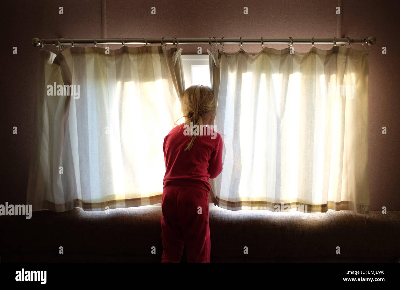 Un soñoliento joven en pijama abre las cortinas para mirar fuera de la ventana en el inicio de un nuevo día Foto de stock