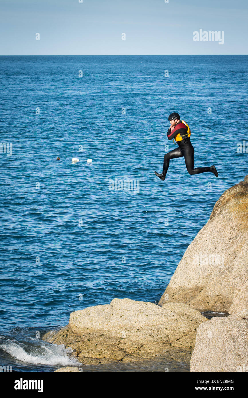 Hombre en traje de neopreno, casco y chaleco salvavidas saltando en las rocas en el mar practicando uno de los deportes coasteering. Foto de stock