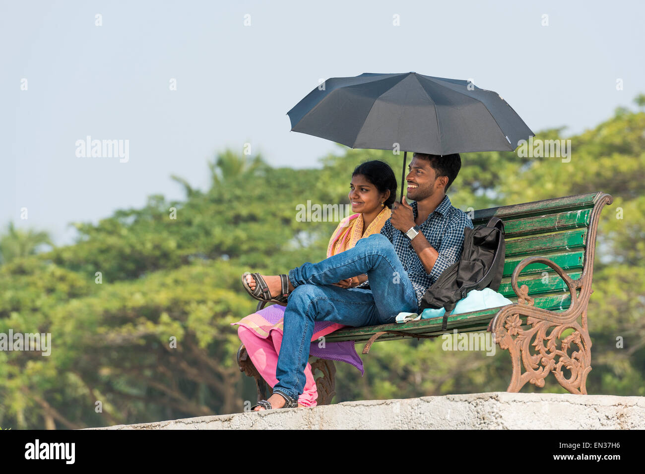 Pareja joven con una sombrilla sentado en una banca del parque, el fuerte Kochi, Kerala, India Foto de stock