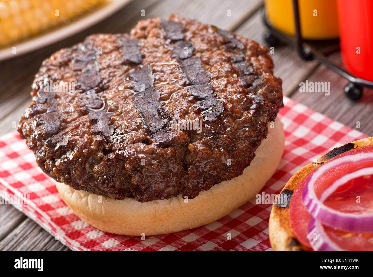 Una jugosa hamburguesa a la brasa con grill marcas en una rústica mesa de picnic con tomate, cebolla, mostaza, ketchup, y el maíz. Foto de stock