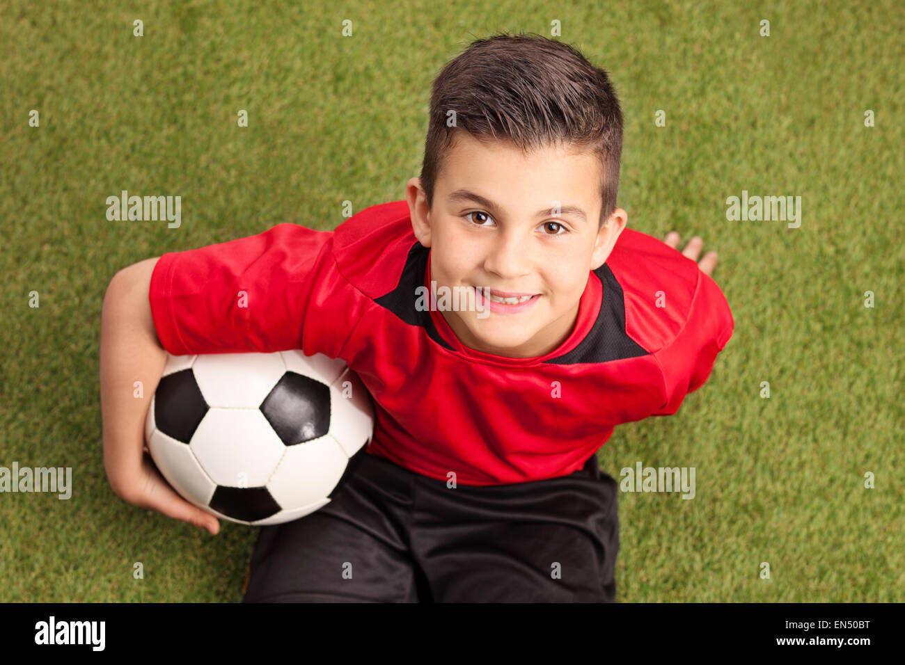 Un alto ángulo de disparo de un jugador de fútbol junior en una camiseta roja sentada sobre la hierba y sonriente sosteniendo un balón Foto de stock