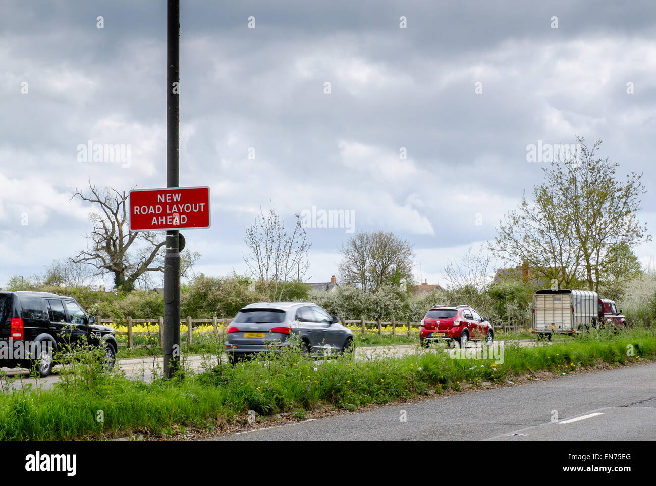 Un "Nuevo diseño de carretera delante" signo en una carretera cerca de un cruce concurrido en las afueras de Cirencester con tráfico en movimiento más allá de Foto de stock