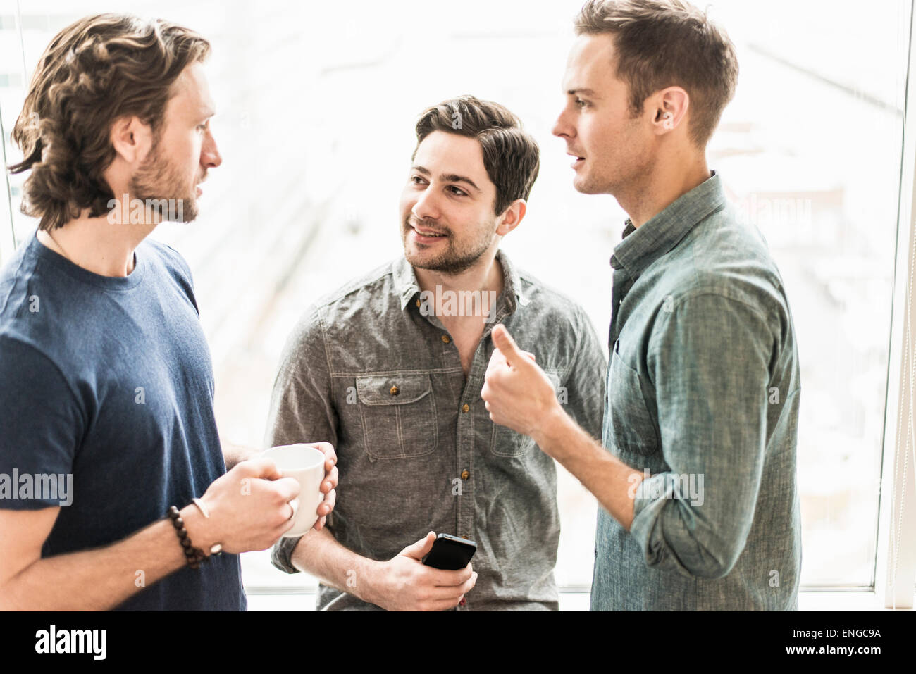 Tres hombres de pie hablando, con una taza de café, uno con un teléfono inteligente. Foto de stock