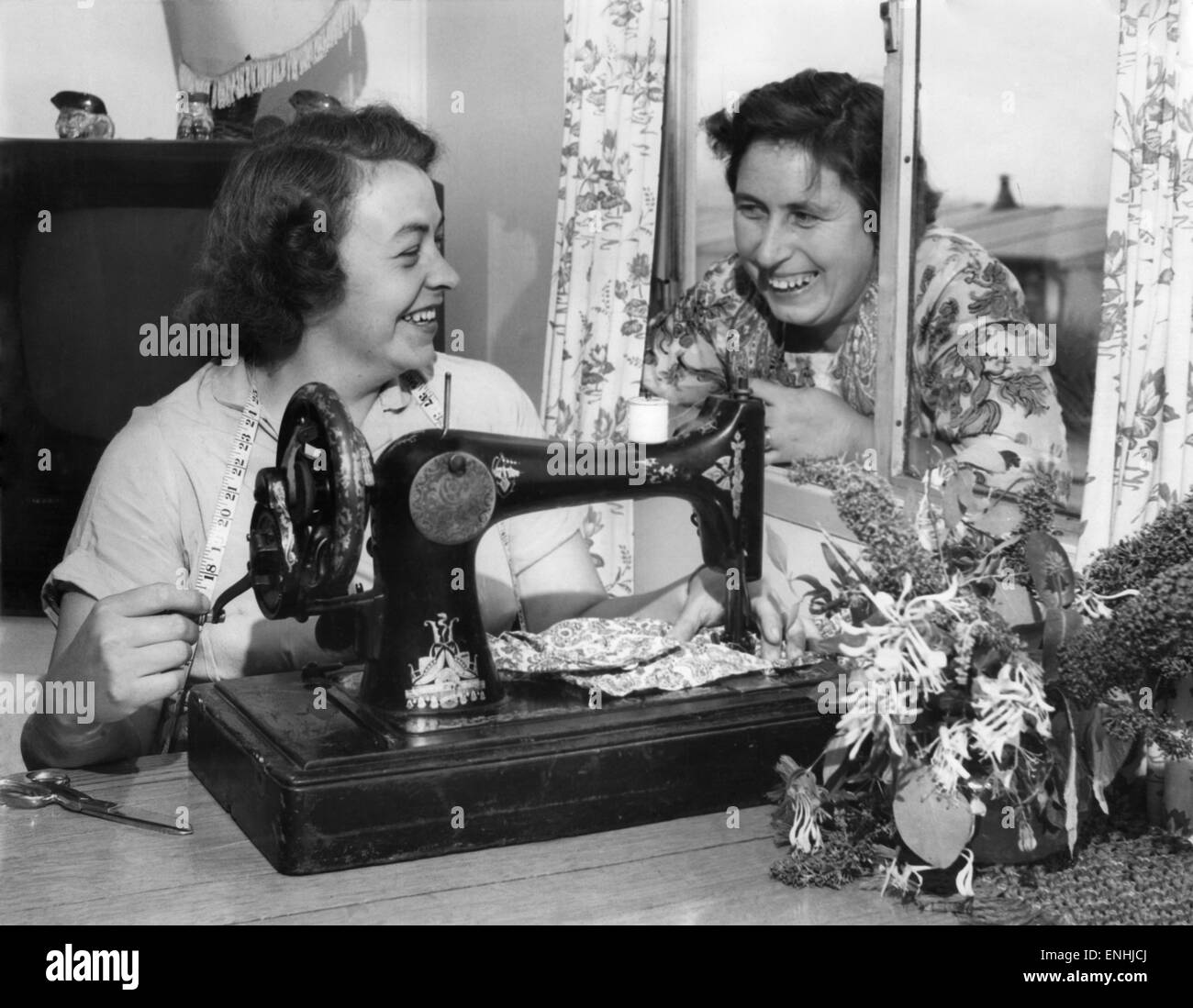 De acuerdo con salud Diverso Ocupado en su máquina de coser es la Sra. Beaty Evans, su vecina, la Sra.  Betty Davies se ve en la ventana para debatir los últimos trajes de baño.  Julio de 1955