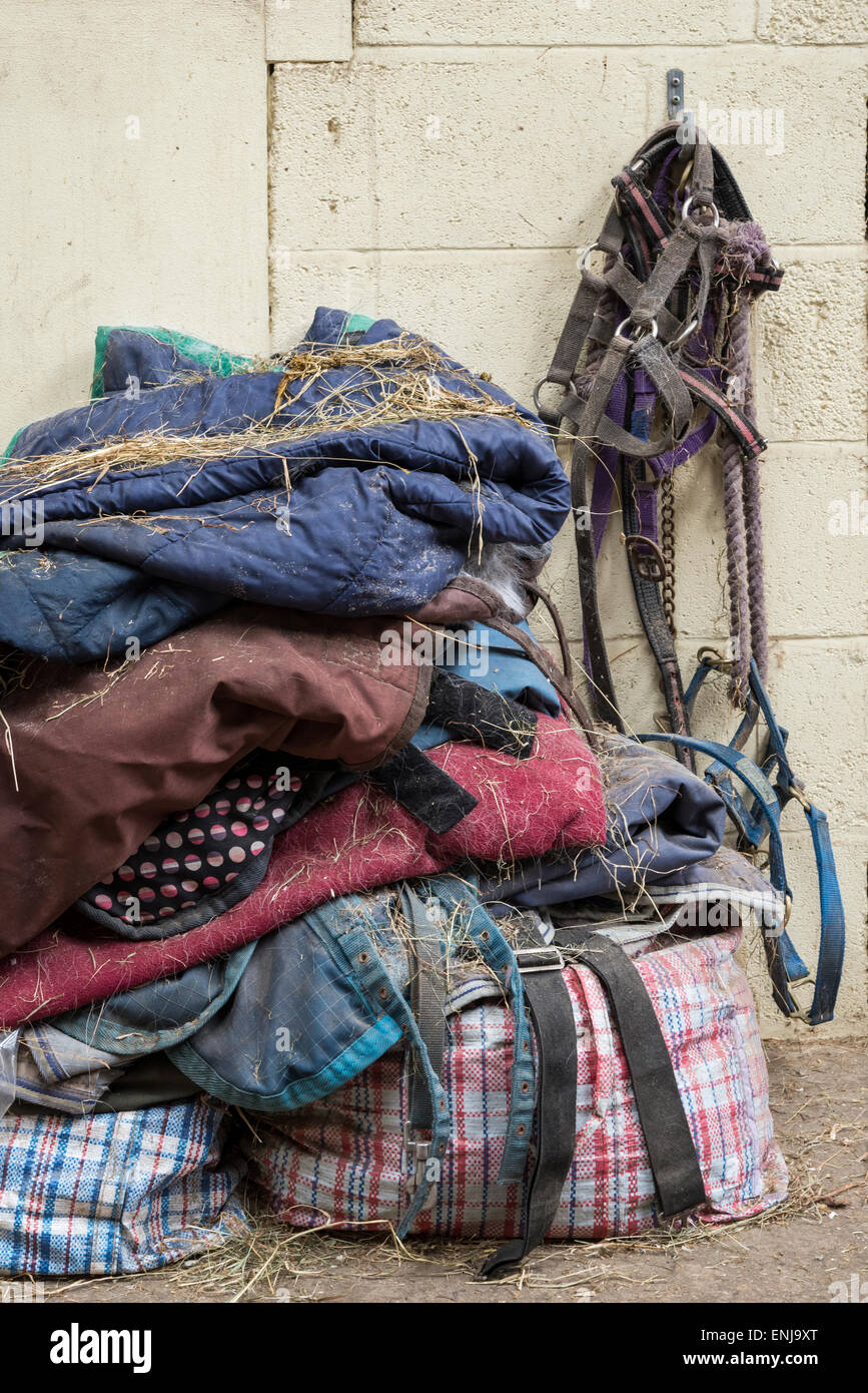 Montón de alfombras y mantas de caballo cubierto de heno con caballos y collares de cabeza llevan cuerdas colgando en un establo. Foto de stock
