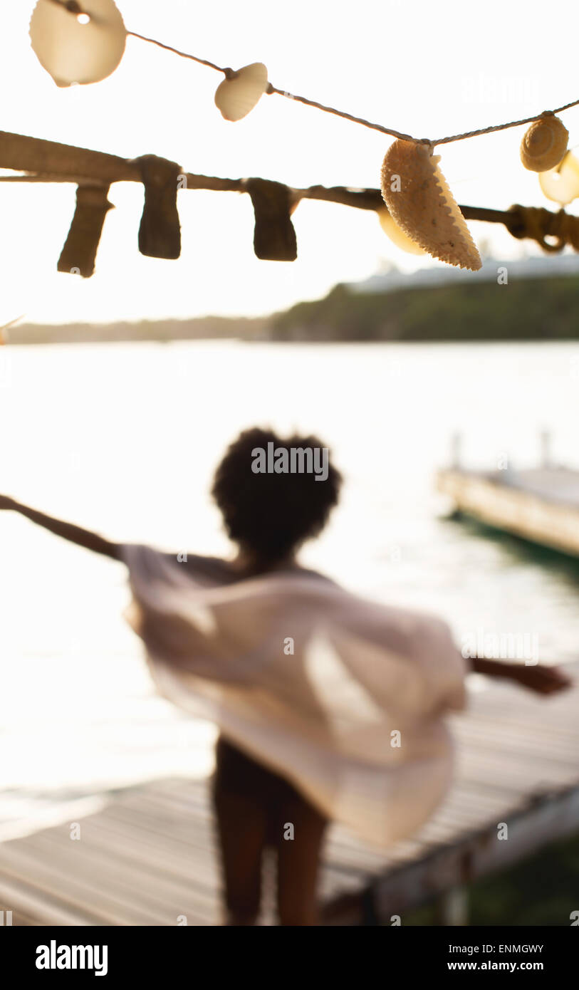 Mujer joven en el embarcadero de madera abrazando un nuevo día, el viento sopla su sarong. Foto de stock