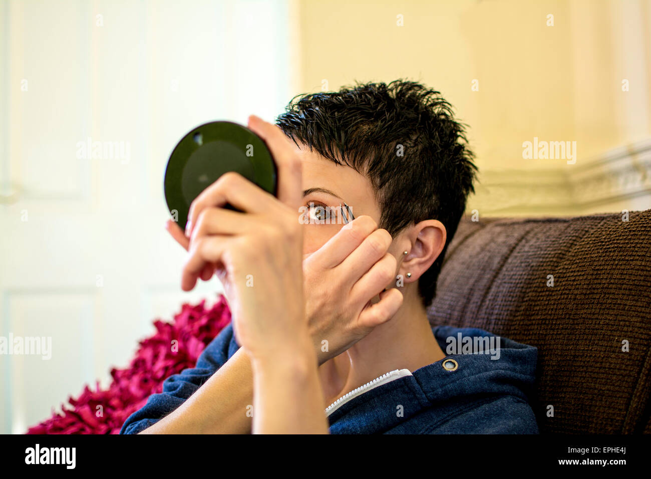 Mujer depilarse las cejas mirando en el espejo, ella se sentó en la sala de estar Foto de stock