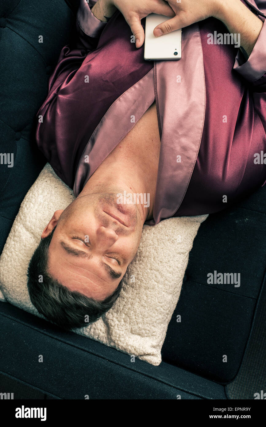 Hombre tumbado y descansando en el sofá en casa. Duerme con la cabeza en una almohada y la celebración de un teléfono móvil en sus manos. Foto de stock