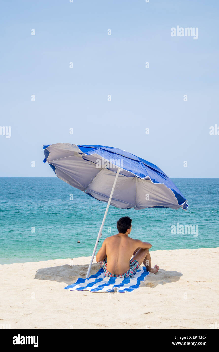 Athletic hombres sentados en una toalla bajo un paraguas azul y gris en la  playa con el océano en el fondo Fotografía de stock - Alamy