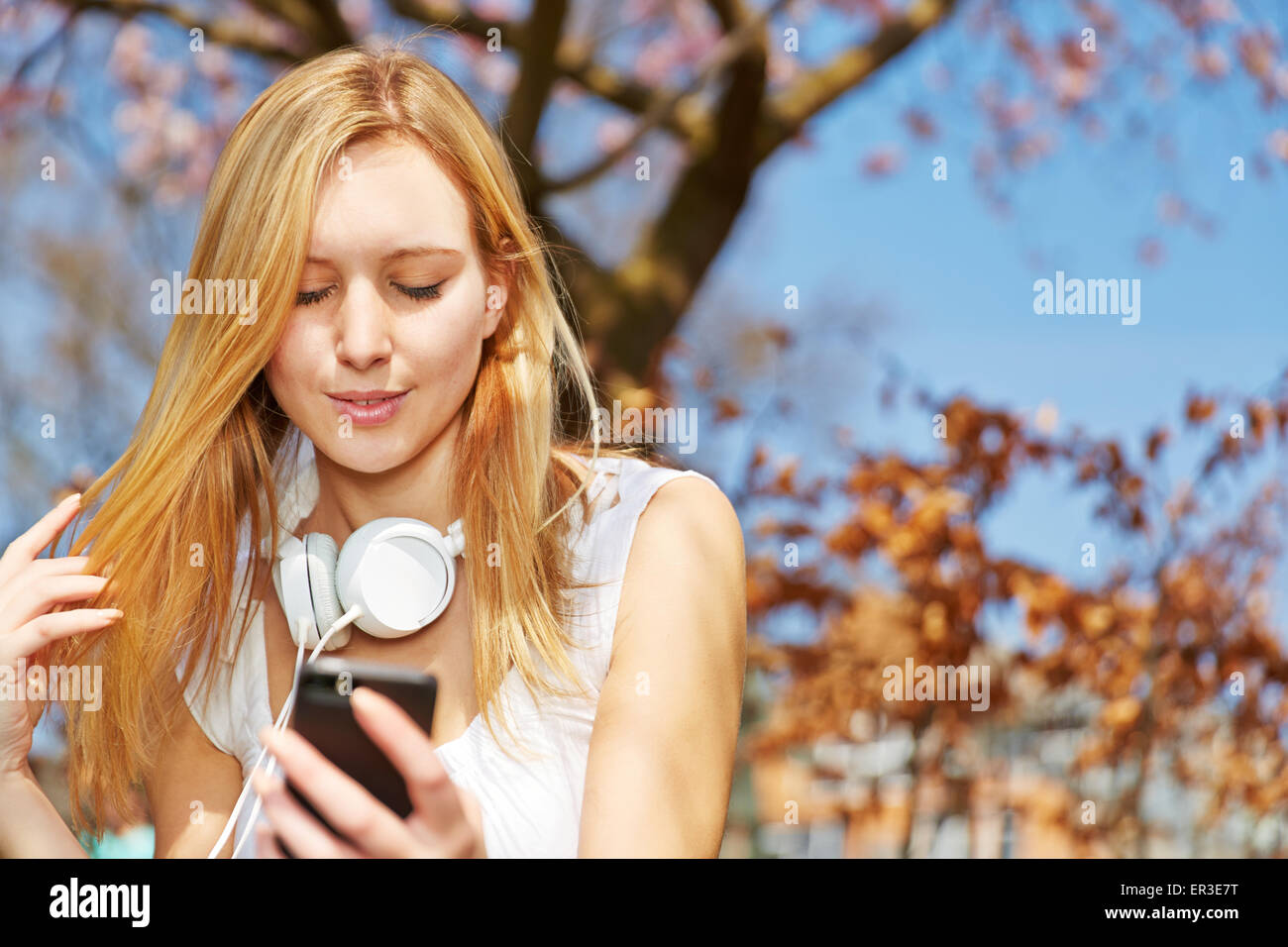 Rubia adolescente sentado con el smartphone y los auriculares en un parque Foto de stock