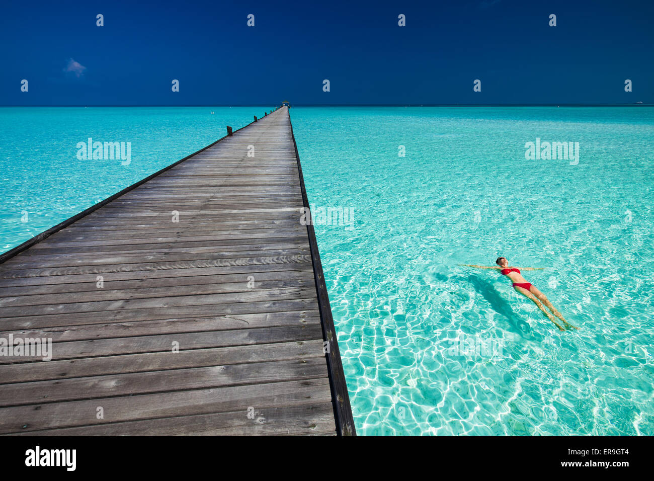 Mujer joven en bikini rojo nadando junto al embarcadero en el agua azul de Maldivas Foto de stock