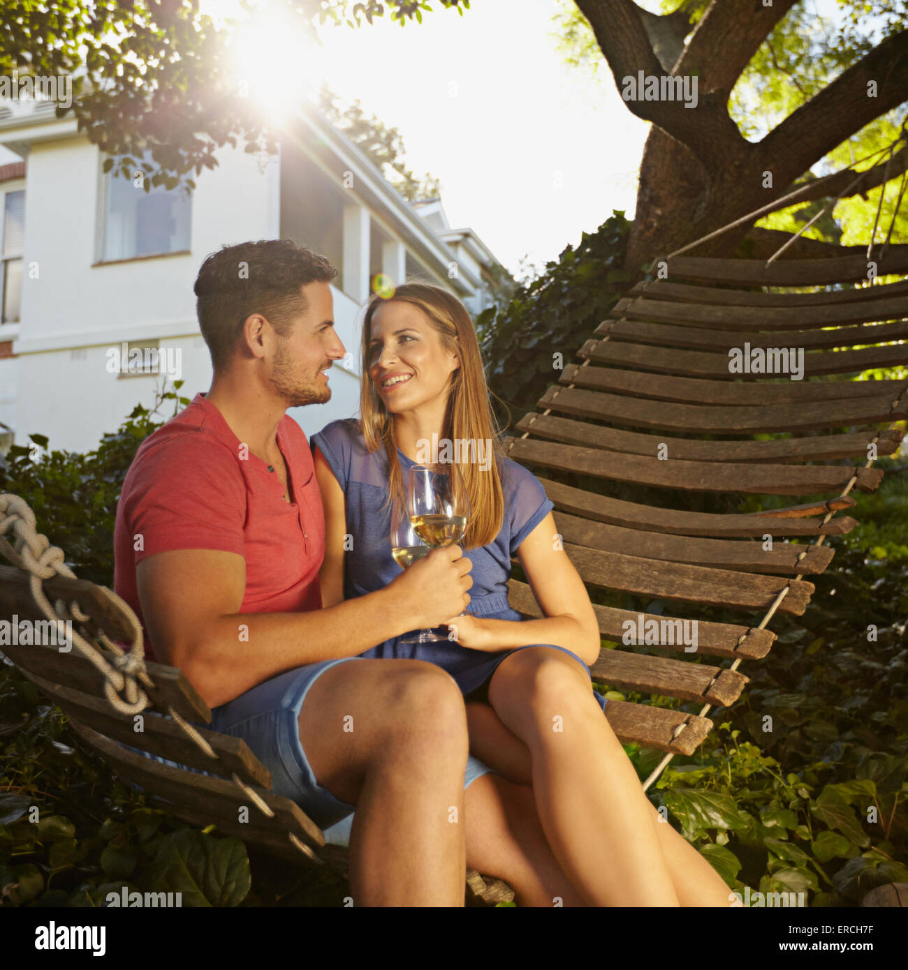 Romántica pareja joven vino tostado mientras está sentado en una hamaca en el patio. Ellos se miran sonrientes y beber Foto de stock
