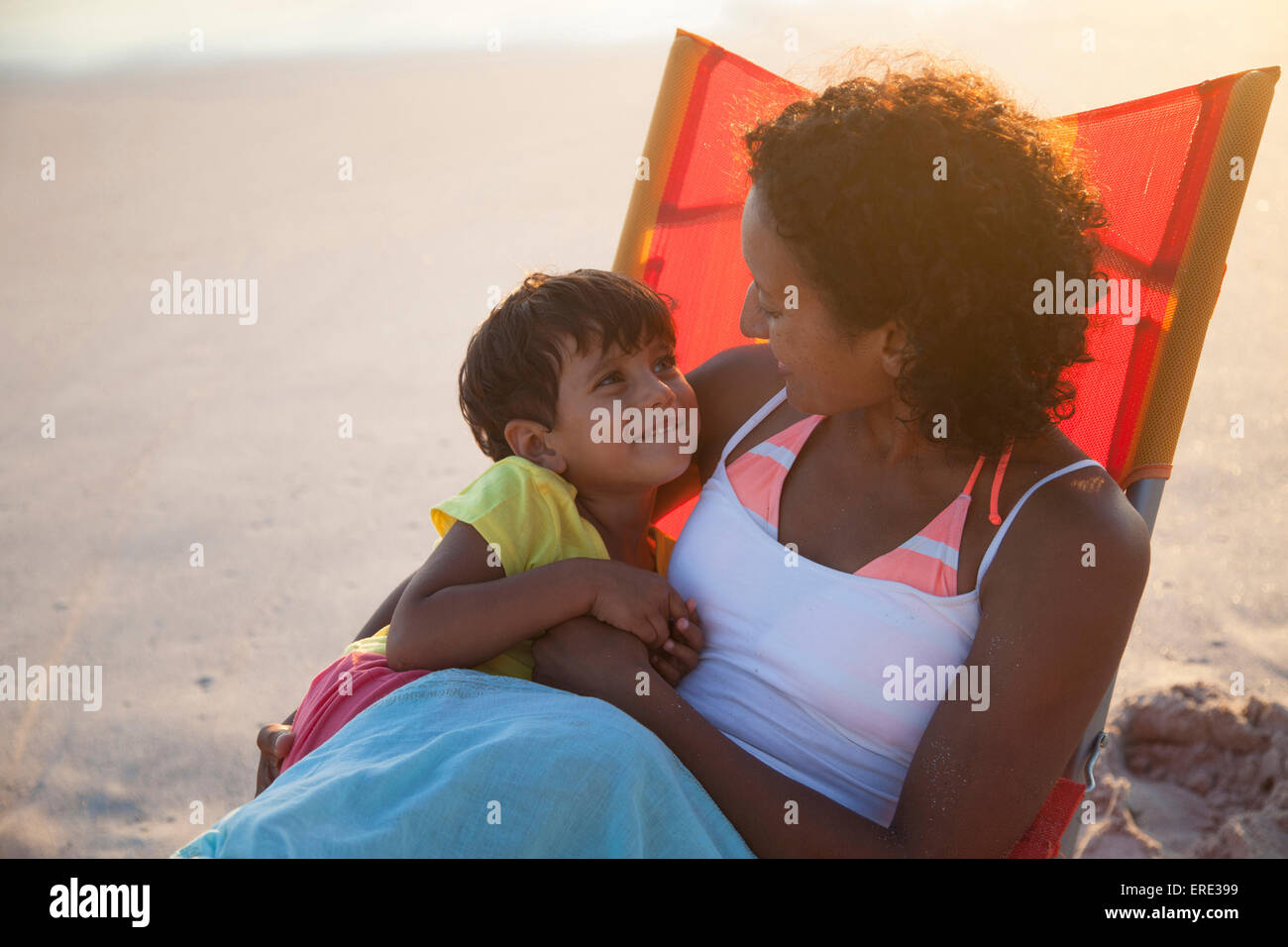 Madre e hija de raza mixta abrazando en una silla en la playa Foto de stock