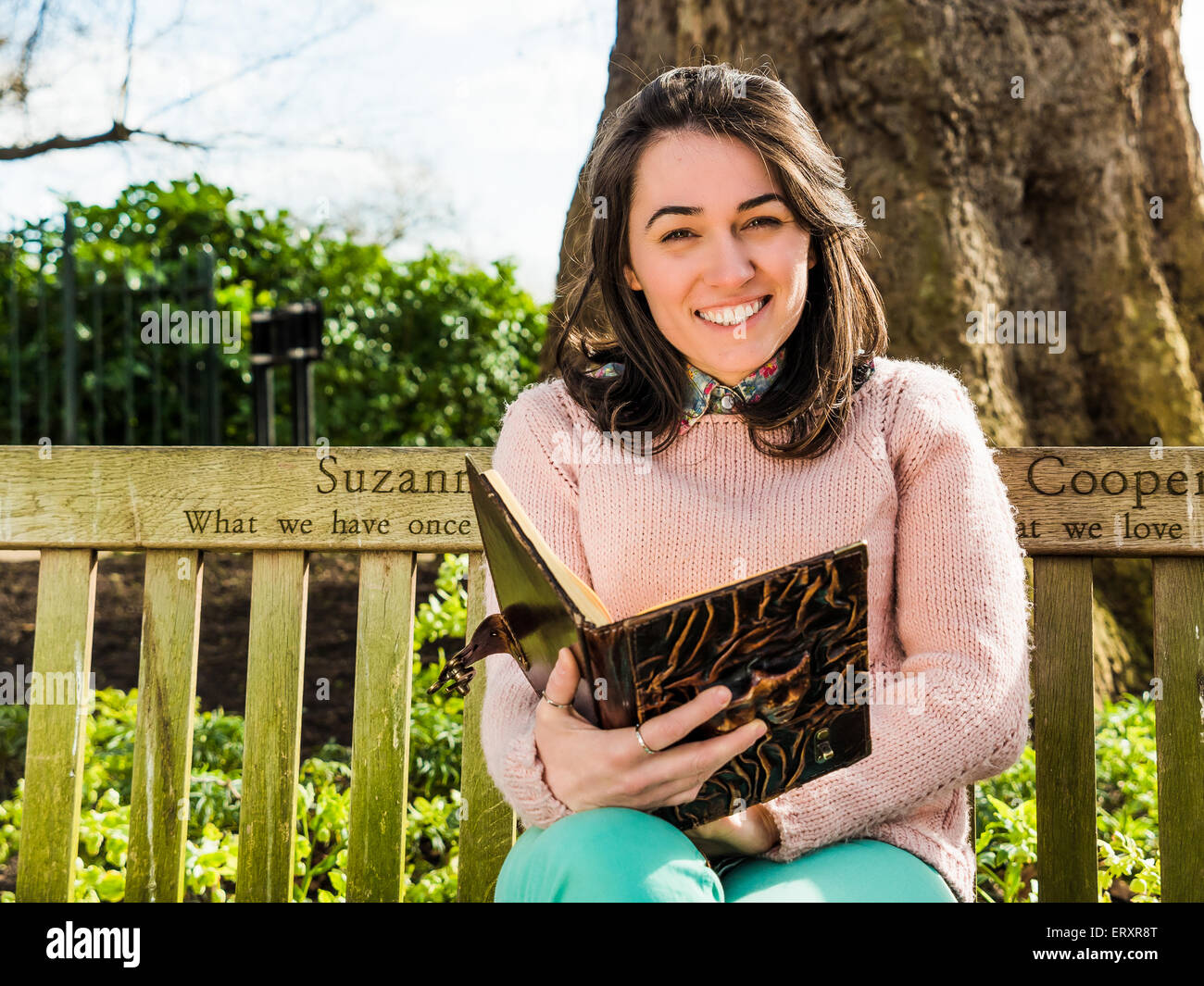 Belleza joven mujer sosteniendo Reserve y sonriente sentado en un banco del parque mirando la cámara Foto de stock