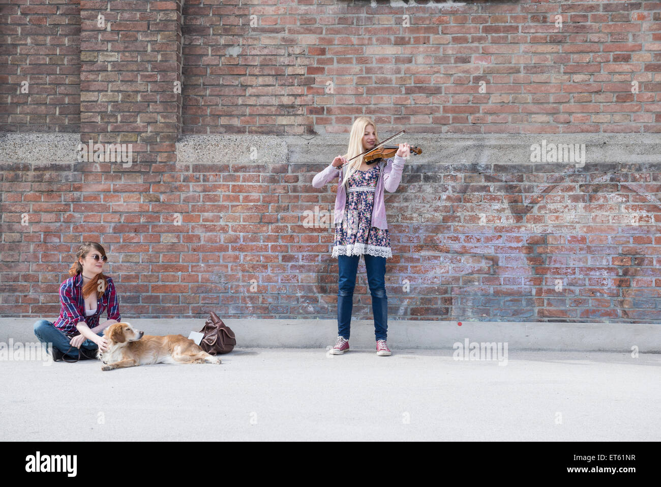 Mujer sentada con su amiga a tocar el violín en frente de la pared de ladrillo, Munich, Baviera, Alemania Foto de stock