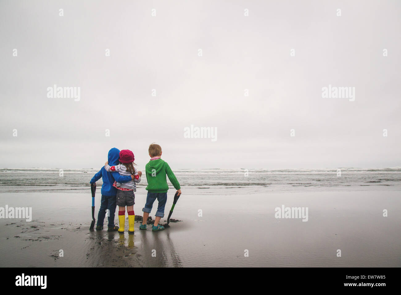 Vista trasera de tres niños de pie en la playa con palas Foto de stock