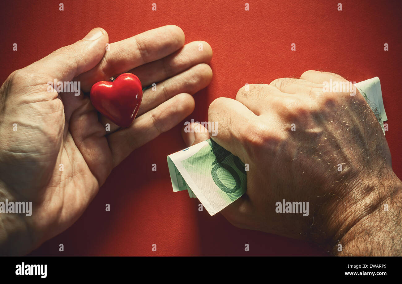 Composición conceptual sobre el amor o el dinero, dos manos masculinas, uno tiene dinero fuertemente y otro rojo corazón como símbolo del amor. Foto de stock