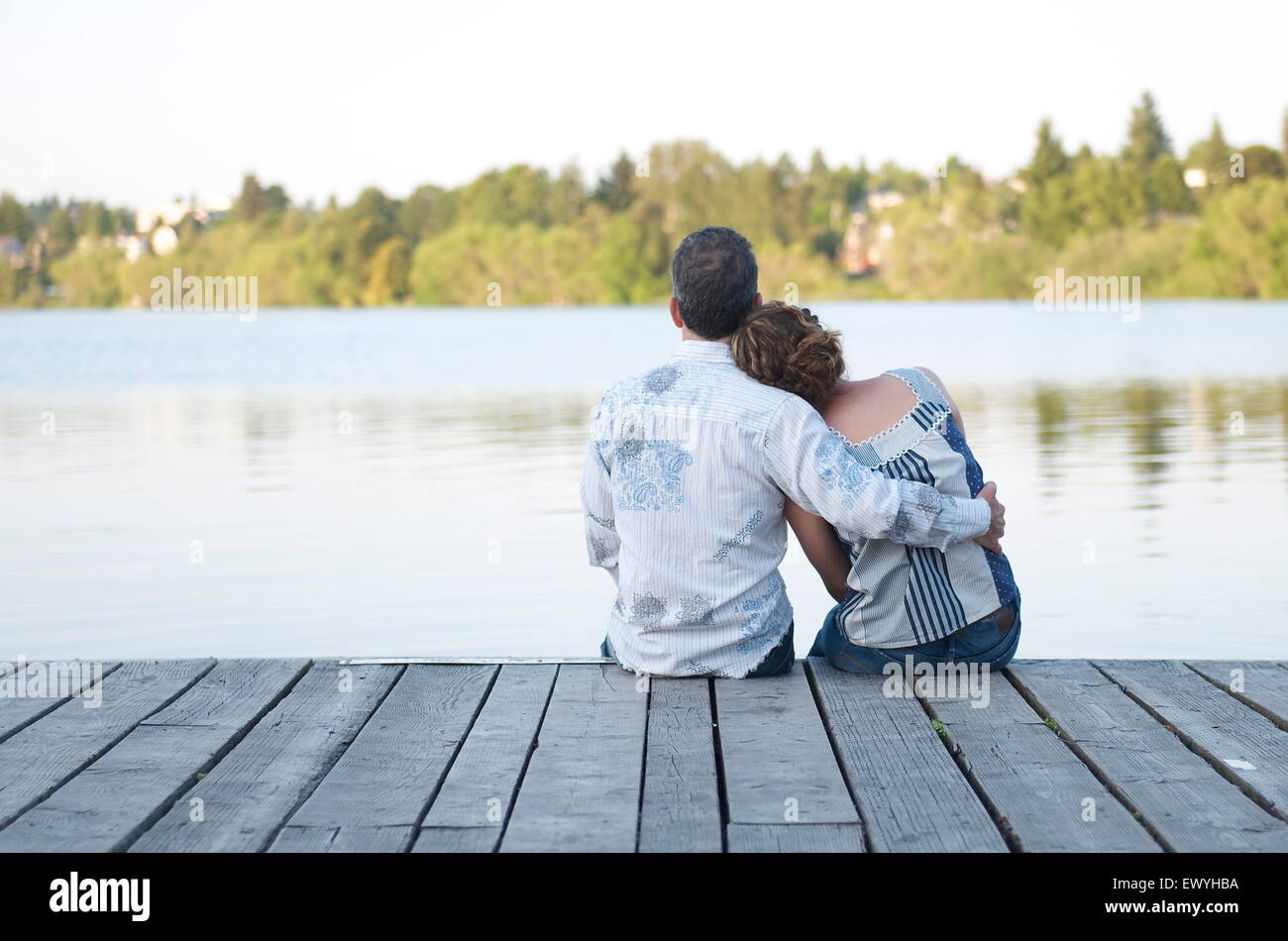 Vista trasera de una pareja sentada en un muelle de madera abrazando Foto de stock