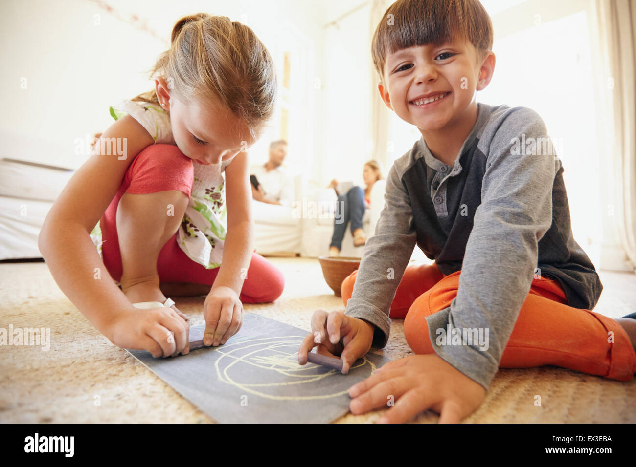 Filmación en interiores de poco feliz niños dibujar y colorear sentados en el suelo con los padres en segundo plano en casa Foto de stock