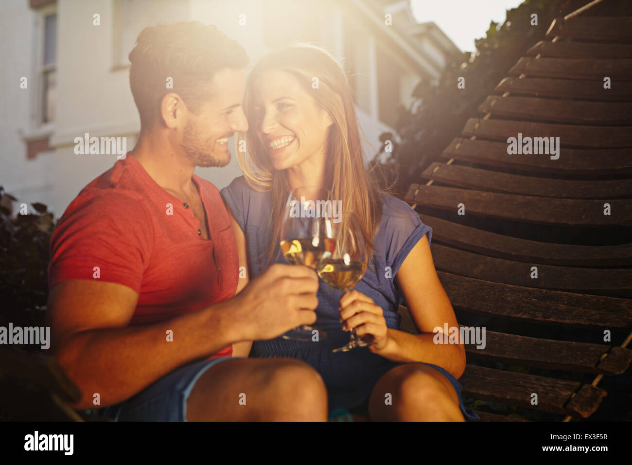 Feliz pareja joven sentado en una hamaca vino tostado y miran sonrientes. Joven pareja amorosa con una copa relajante Foto de stock