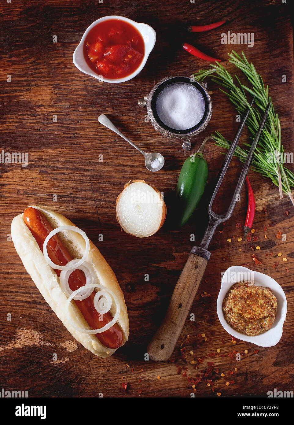 Perro caliente casero con ingredientes mostaza, salsa de tomate, cebolla, pimiento, Romero. Sobre la mesa de madera con carne tenedor y cuchara Foto de stock