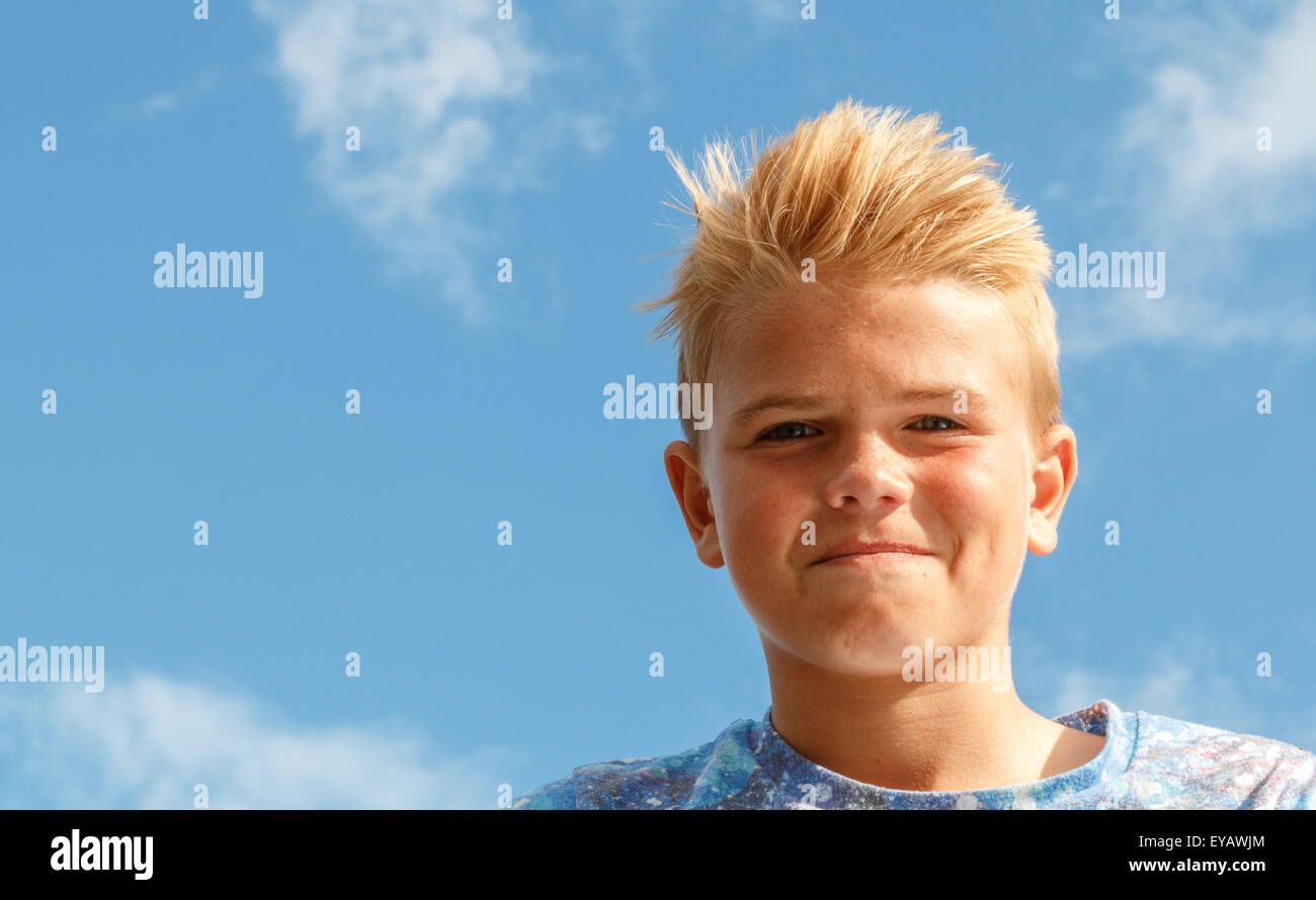 Rubia adolescente (13 años) con pelo de punta smirking o sonriendo mirando cool contra un brillante cielo azul con nubes blancas Foto de stock
